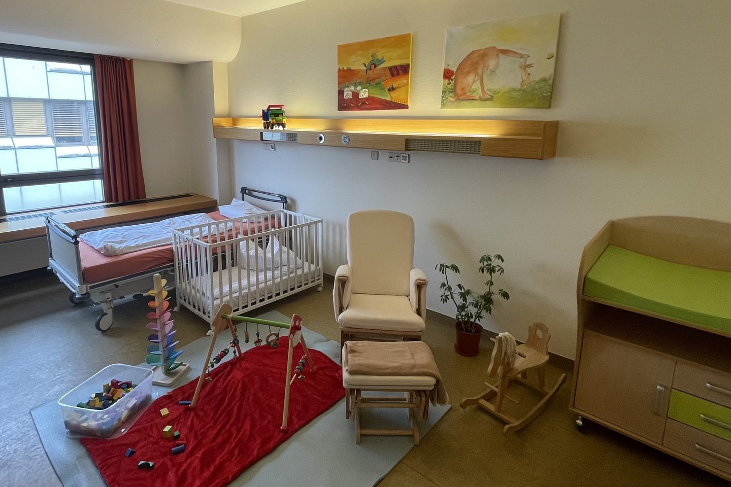 Patientenzimmer mit Bett, Kinderbett, Wickelkommode, Spielsachen, Sessel und kindgerechte Bilder