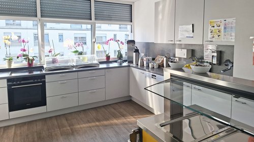 Eine weiße Küche in L-Form mit Herd, Backofen, Geschirrspüler, Spüle, Kühlschrank, usw. Im Hintergrund sind drei Fenster mit Blumen auf dem Fensterbrett. Das mittlere Fenster ist gekippt.