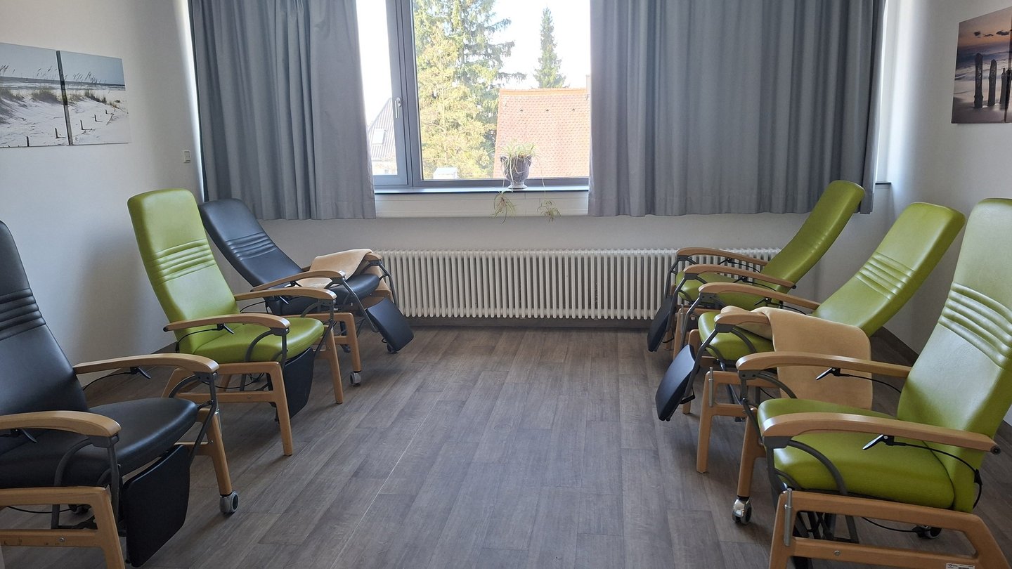 Raum mit sechs Liegen, jeweils drei stehen sich gegenüber. Auf zwei Liegen sind Decken. Im Hintergrund ist ein Fenster mit halb zugezogenem dunklen Vorhang.