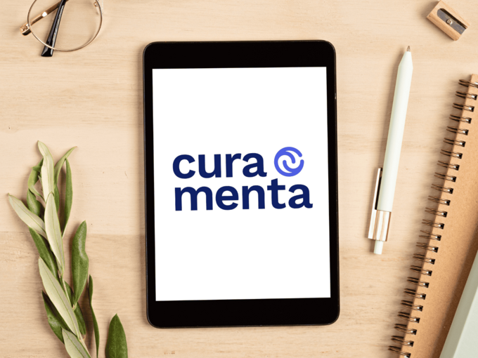 Schreibtisch mit Blöcke, Stifte, Brille und Tablet auf dem das Curamenta-Logo zu sehen ist.