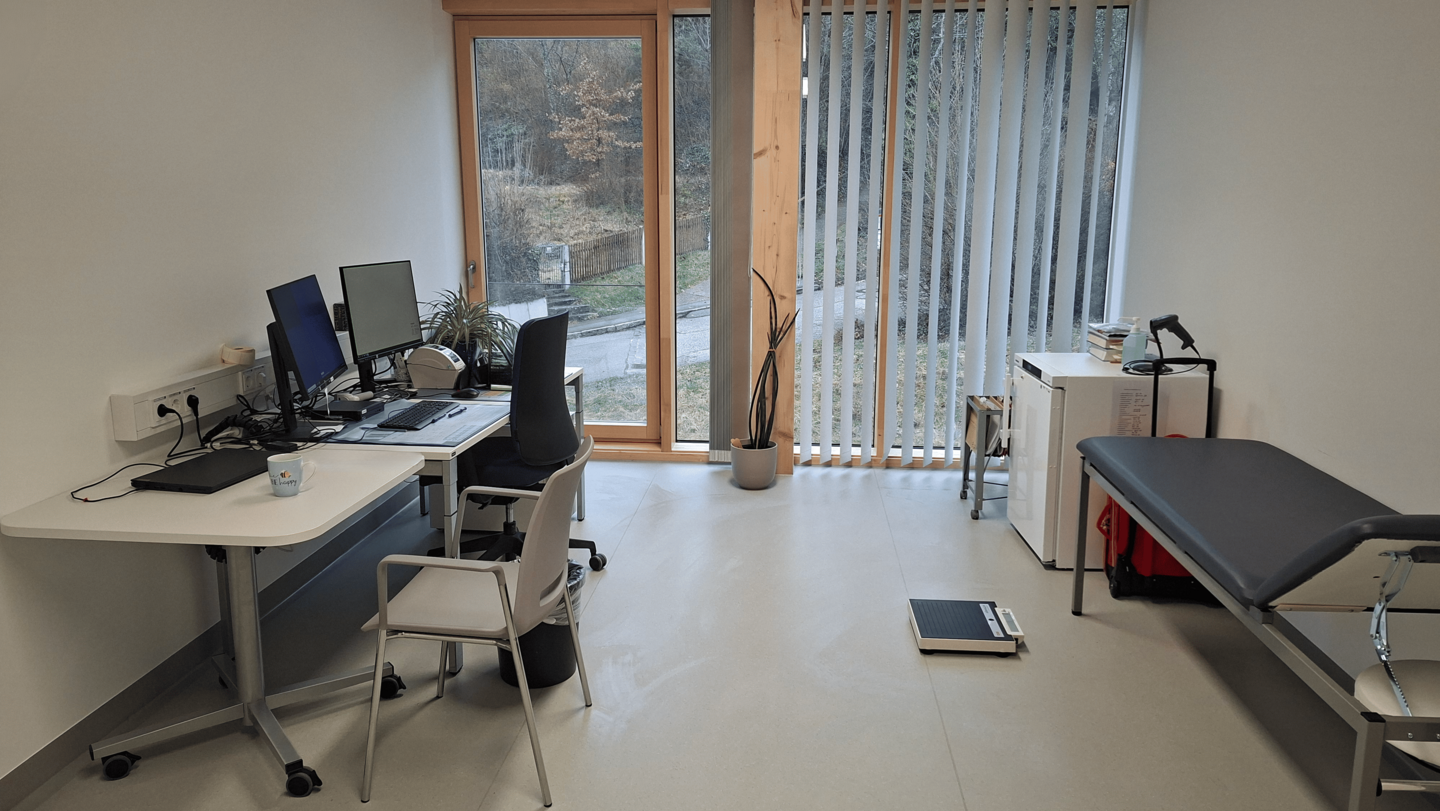 Behandlungsraum mit Schreibtisch mit zwei Bildschirmen, bodentiefe Fenster, eine Waage und eine Behandlungsliege